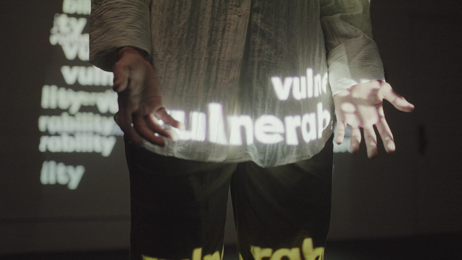 The lower midsection and upper thighs of person is seen. This person is wearing a white button up shirt, the word 'vulnerability' is projected repeatedly over them, and a screen behind them also shows this projection.