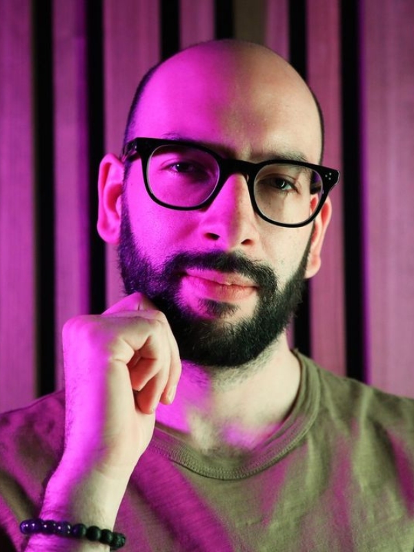 Shoulder-and-up headshot of Connor. He is white, male, with closely trimmed and shaped beard, and a near-shaven head. He wears thick black rimmed glasses, a beaded bracelet, and a dark mustard-green shirt. The lighting is bold pink on his right half, while left half is more natural white-yellow. One hand is held up against the side of his chin as if in mid-thought, and his eyebrows and eyes betray a mix of curiosity and inner reflection.