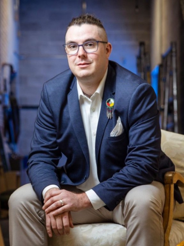 Deneh is seen sitting on a cream-coloured chair. He is slightly bent forward with his elbows resting on his thighs. He is wearing a blue blazer, white button up shirt, and beige/cream pants. In the background you see blurred grey brick walls. Deneh is wearing glasses with blue-ish frames. He has a beaded dreamcatcher-looking pin on the collar of his blazer.