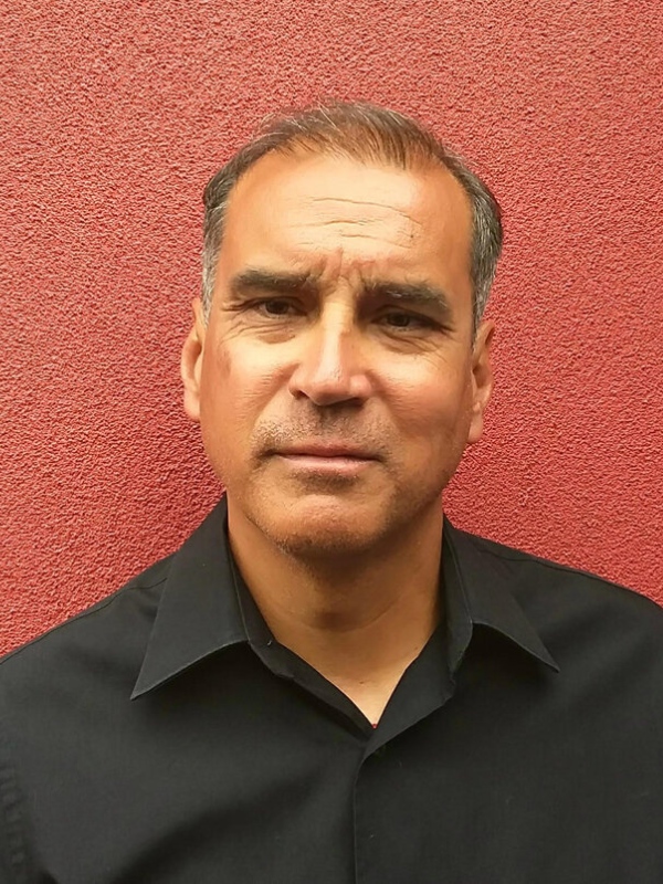 Dr. Dwayne is seen up against a red stucco wall. He is wearing a black collared shirt.