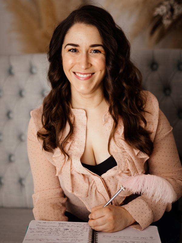 Elisa sits on a grey sofa, smiling at the camera. Her long brown hair is curled. She is wearing alight pink blouse with tiny black dots on it. The collar is ruffled. She is holding a pen and a notebook sits on her lap.