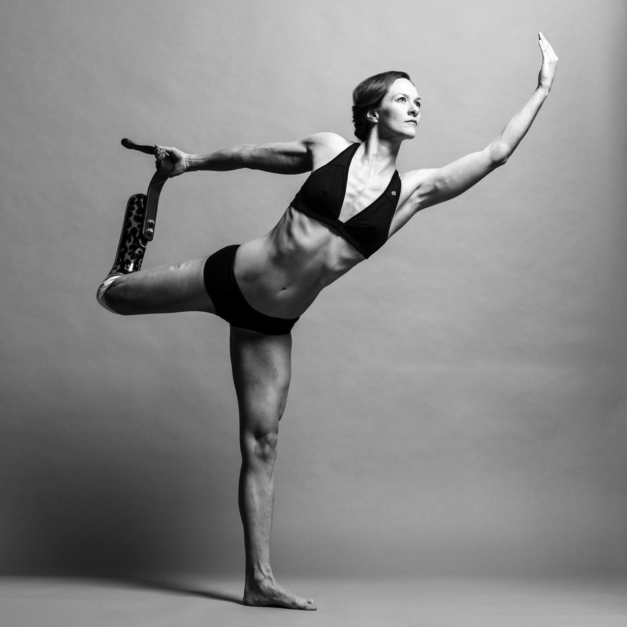 A powerful photo of Kristi standing on one leg, with her other leg (with her prosthetic) bent back behind her. She holds the base of her prosthetic leg with her right arm. Her left arm is held up in front of her and upwards, this is also the direction her face is looking. She is wearing a black set of clothing - a v-neck sports bra and briefs.