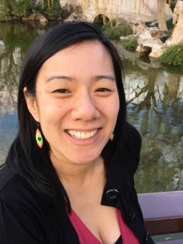 Photo of Lucy Lu, from her chest up. She is smiling at the camera, her dark hair is tucked behind her ears - where you can see her colourful earrings. Her smile is wide. You can see a pond behind her, with reflections of a blue sky, rocks, and trees. She appears to be standing on a walking bridge, with part of the railing visible behind her.