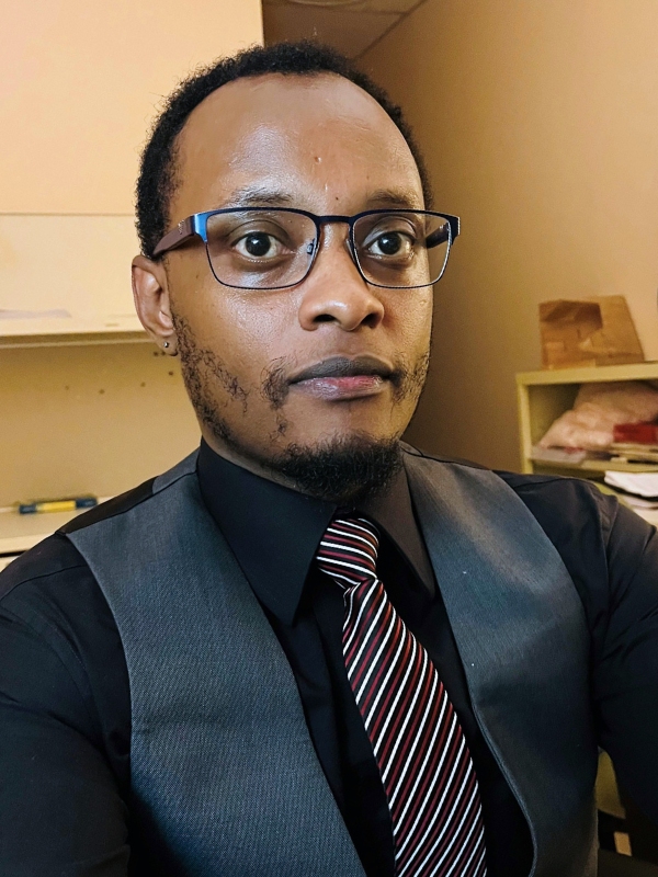 Photo of Mukonzi Musyoki wearing a dark grey vest, black button up dress-shirt, and a black-white-red striped tie. He is looking directly at the camera, wearing dark framed glasses.