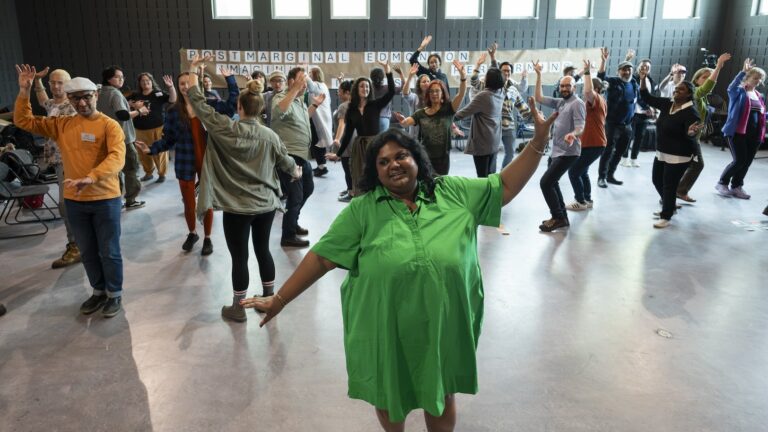 A woman in a green dress with her arms outstretched leads a room full of people who are doing a physical exercise.