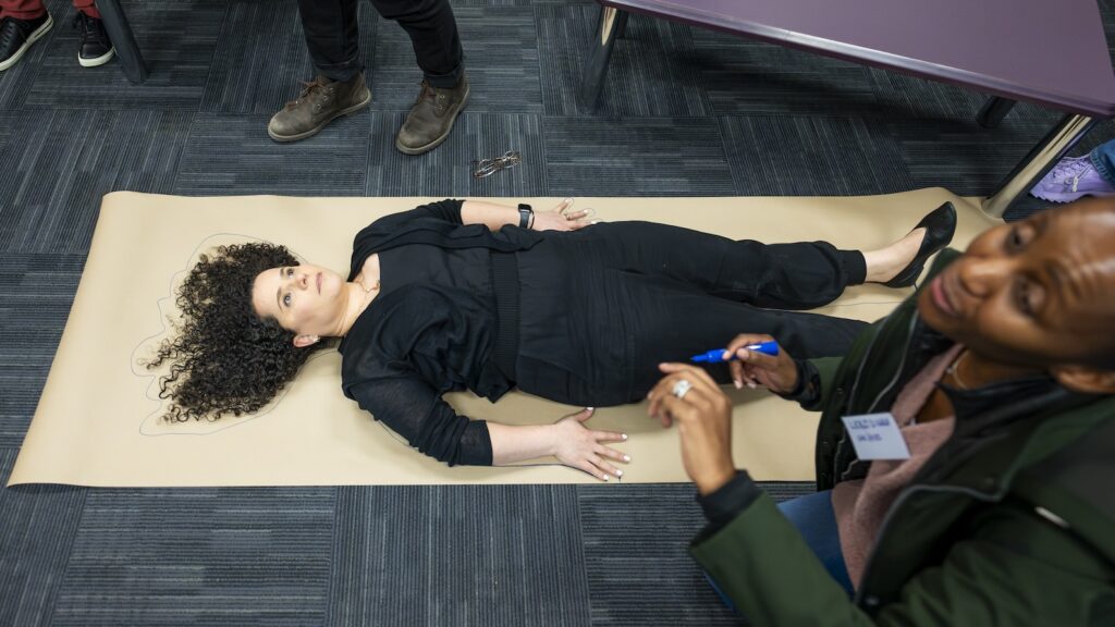 A woman lies on a long brown paper while someone traces her outline.